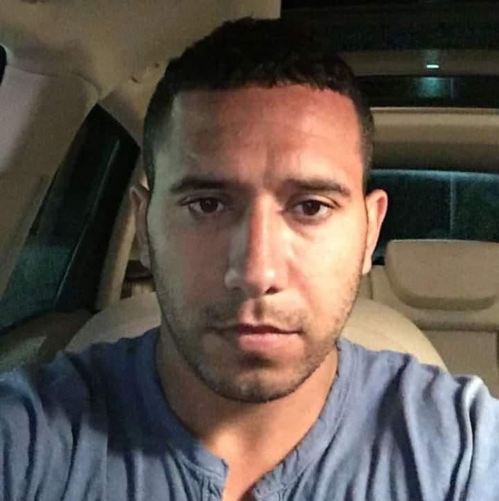 Man with short dark hair and stubble wearing a blue t-shirt inside a car.